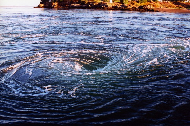 A swirling ocean whirlpool near a rocky shoreline.