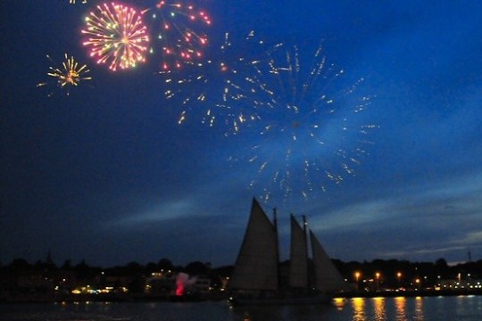 Fireworks over a sailboat at night with city lights in the background.