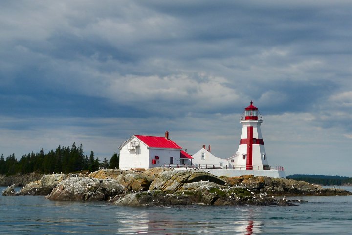 Lighthouse with red roof on rocky island with cloudy sky.