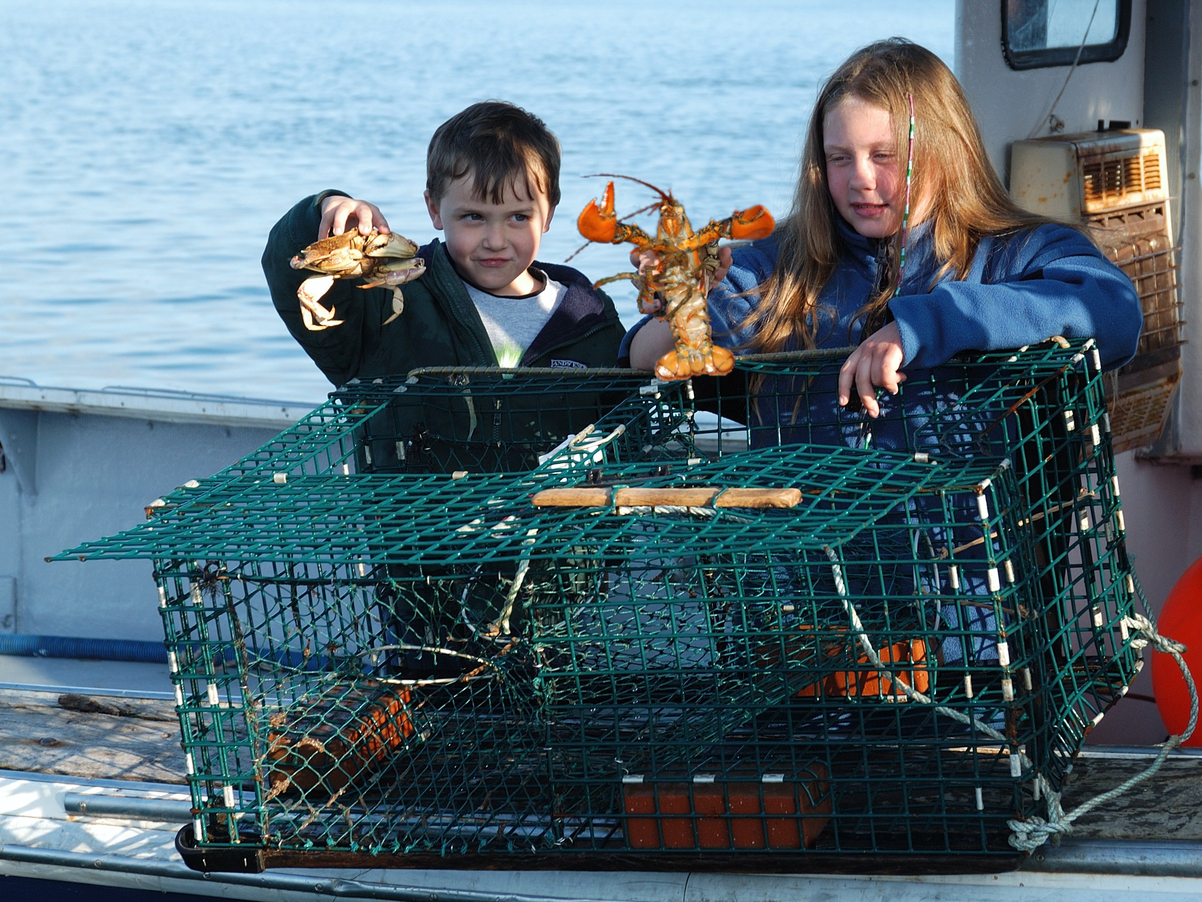 Two children holding a lobster and crab on a boat with a lobster trap.